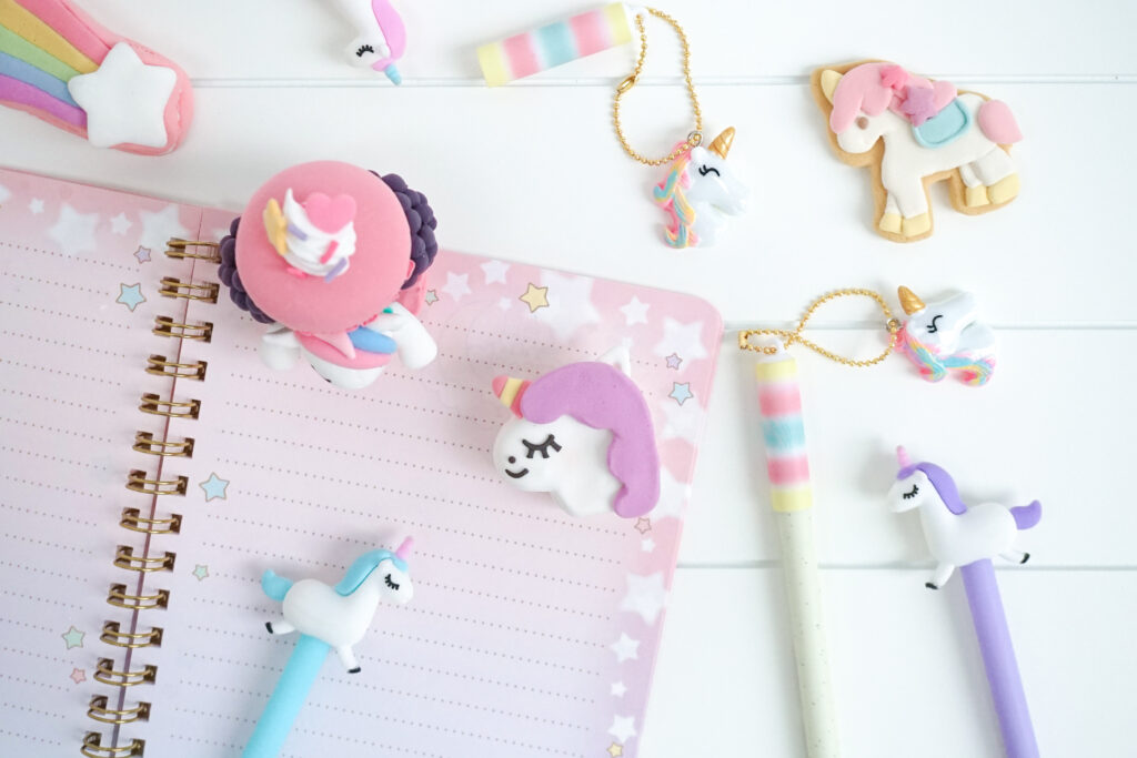 Pastel unicorn-themed stationery on a white surface: pink star notebook, unicorn pens with toppers, and cute charms to decorate a desk.