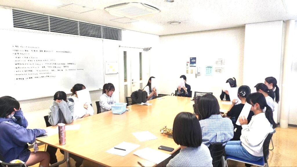 Group of students seated around a long conference table in a classroom, many covering their faces with papers as they read or hide from view.