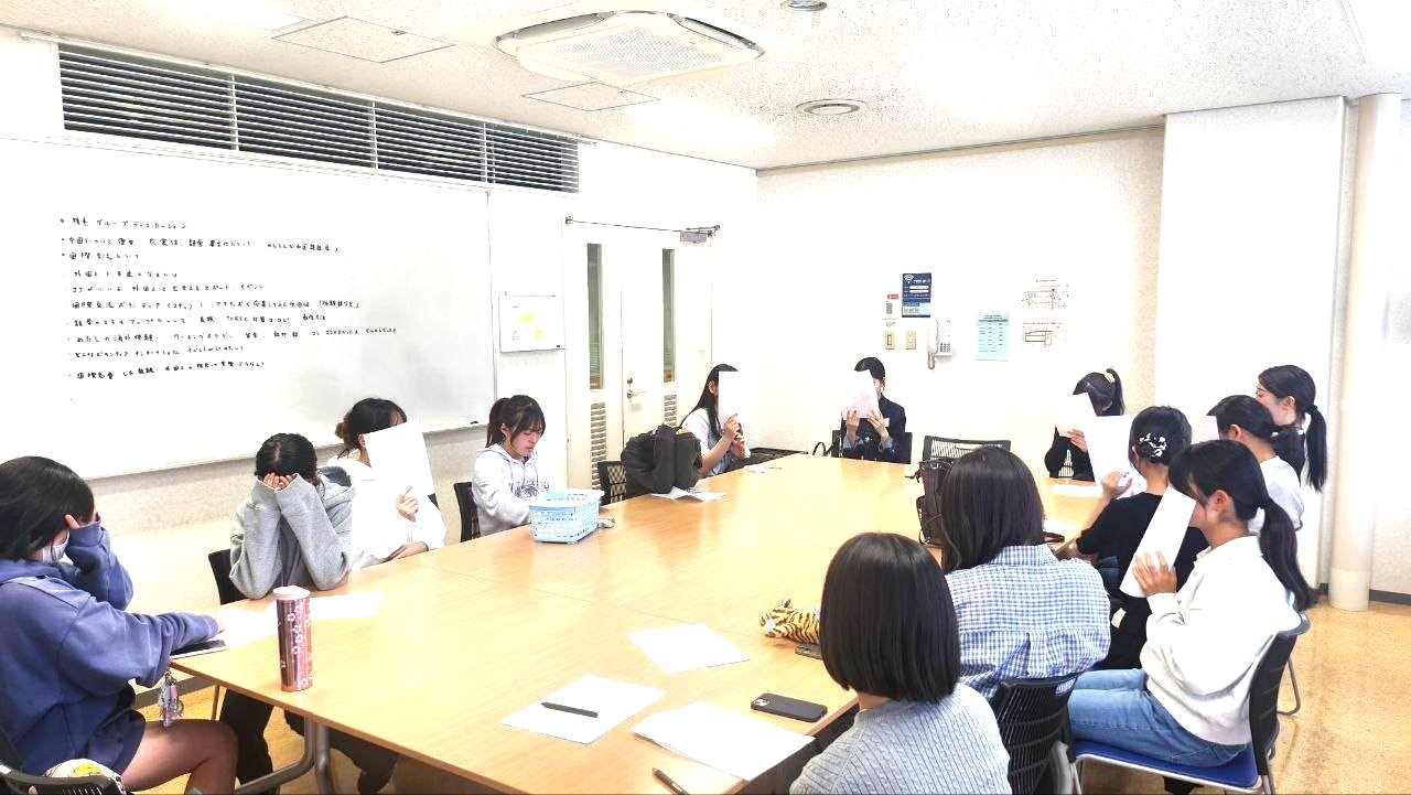 Group of students seated around a long conference table in a classroom, many covering their faces with papers as they read or hide from view.