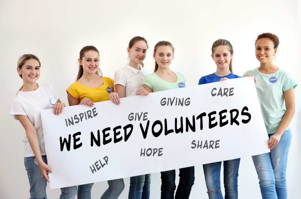 Six smiling young volunteers stand side by side, holding a large banner that reads 'We Need Volunteers' against a white wall.