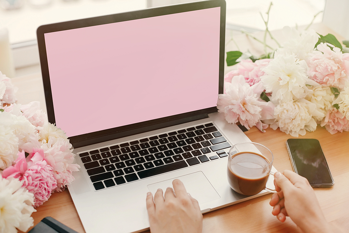 Laptop with a pink screen on a flower-filled desk; hands typing, with a cup of coffee and a smartphone nearby.