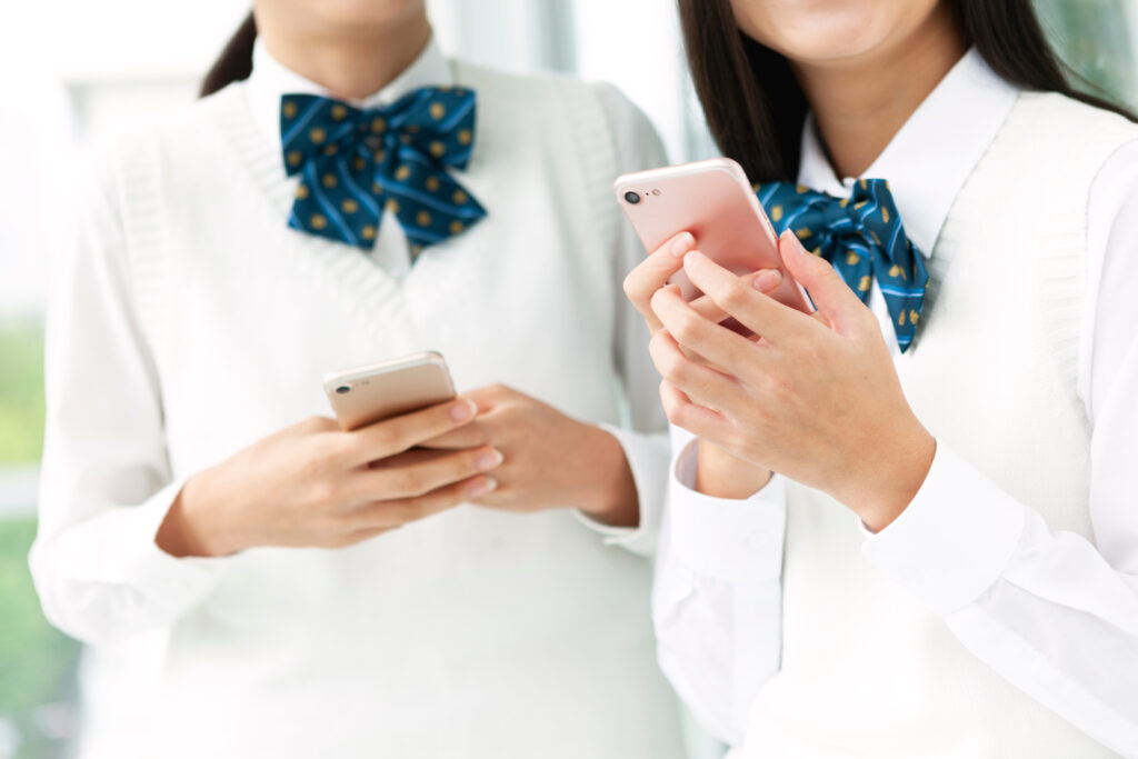Two students in white uniforms with blue patterned bows, looking at their smartphones side by side.