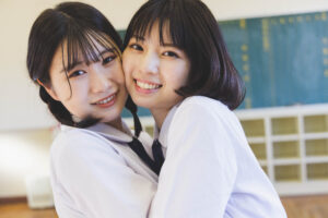 Two smiling students in white uniforms hug each other in a classroom, with a chalkboard in the background.