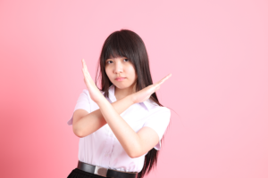 Young woman in a white blouse crosses her arms in an X to signal 'no' or 'stop' against a pink background.