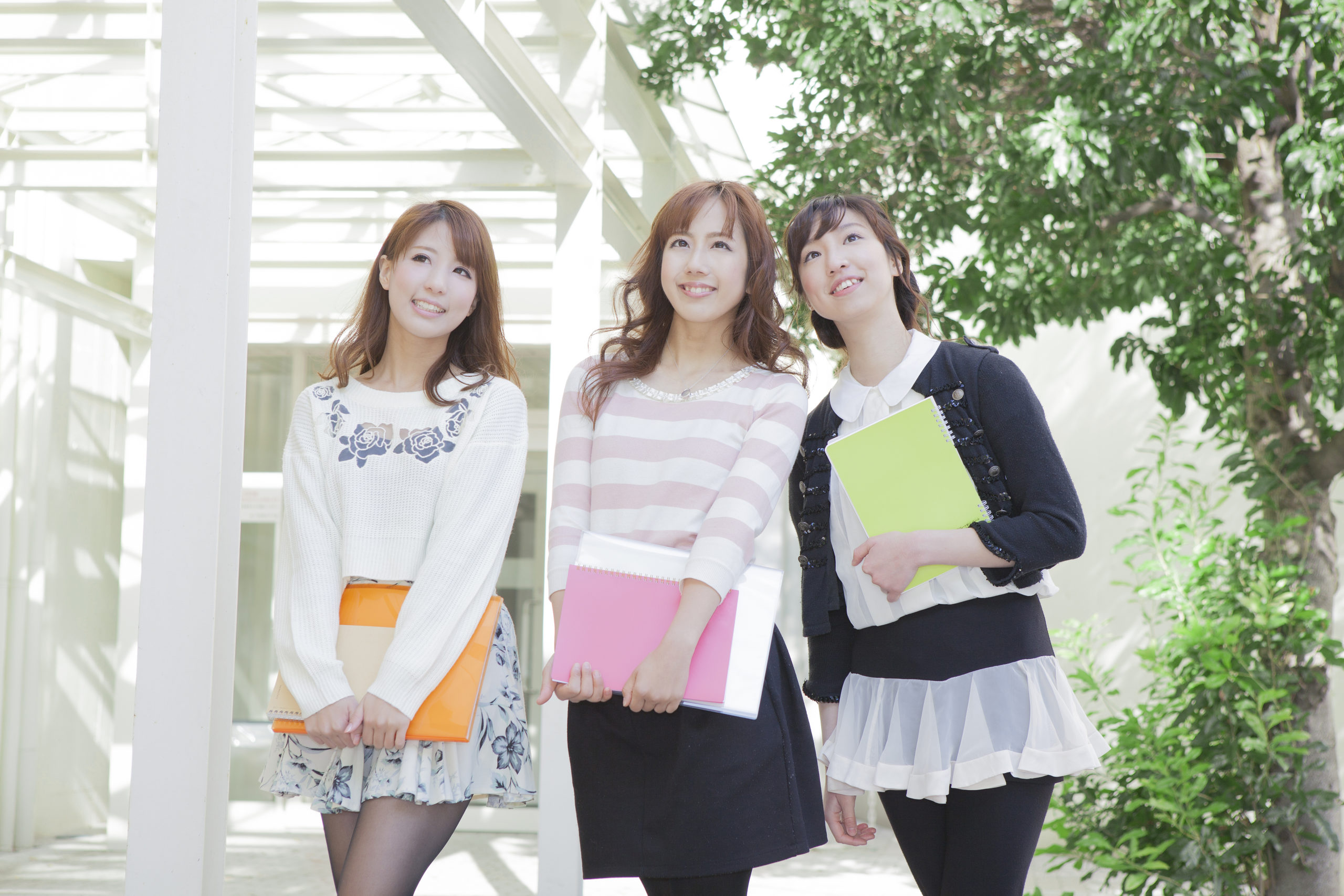 Three young women standing outdoors, smiling and holding colorful notebooks.