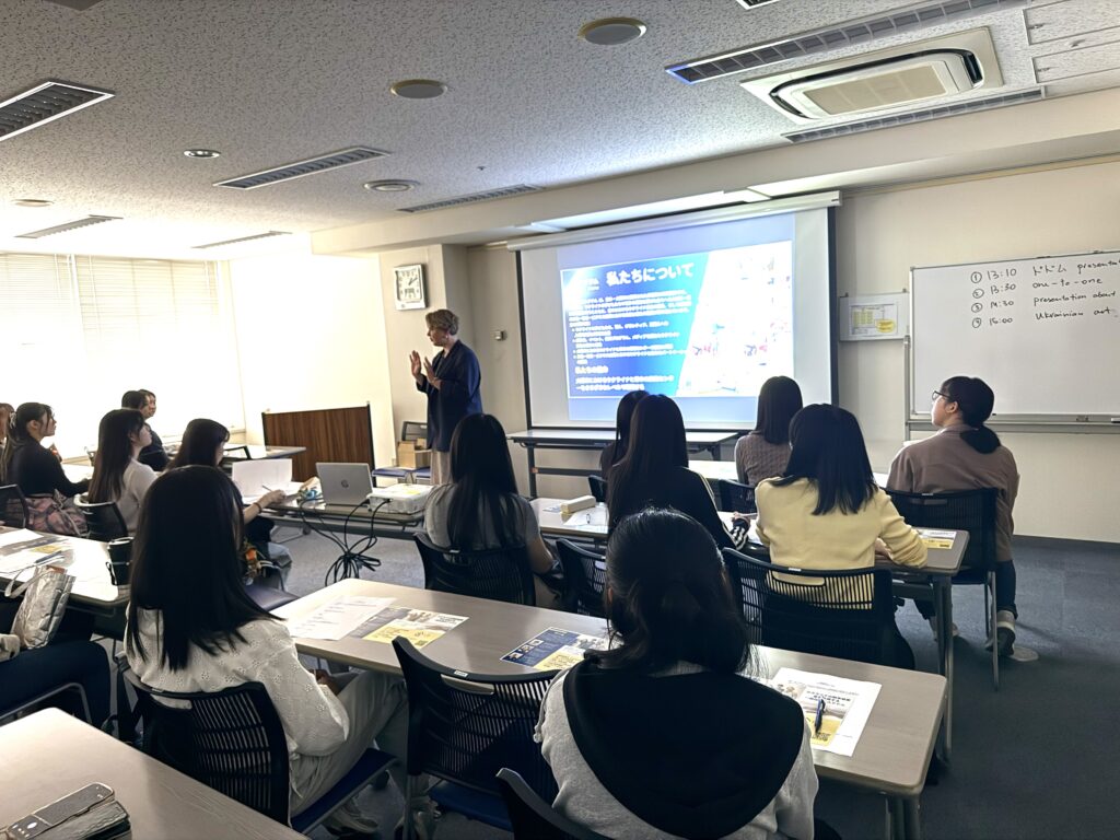 Classroom presentation with a speaker at the front and students seated facing a projected slide fielding their attention to a large screen.