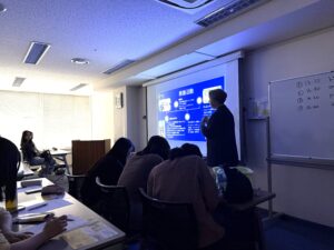 Instructor presenting at the front of a classroom with a blue slide projected on a screen, while students take notes at their desks.
