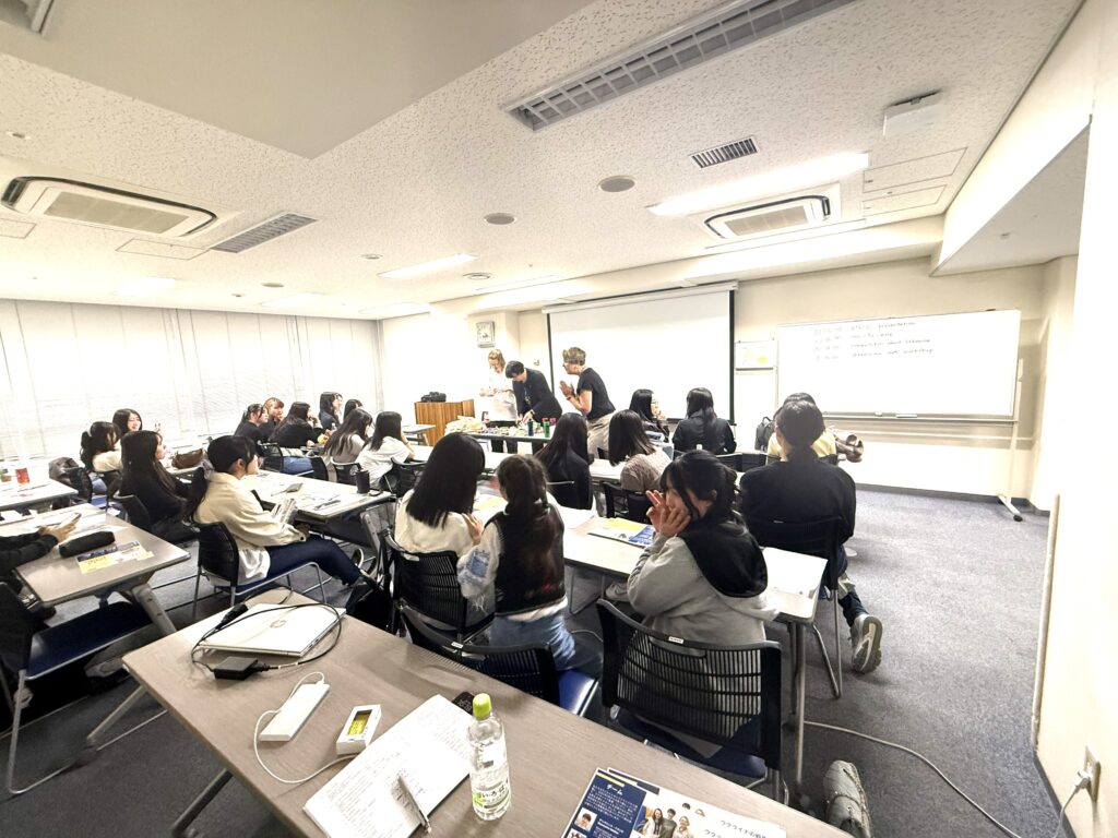 A crowded classroom with students seated at desks facing a presenter at the front and a whiteboard.