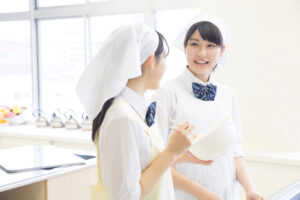Two young women in white chef uniforms with hats share a conversation in a bright kitchen or classroom setting, one holding a clipboard.