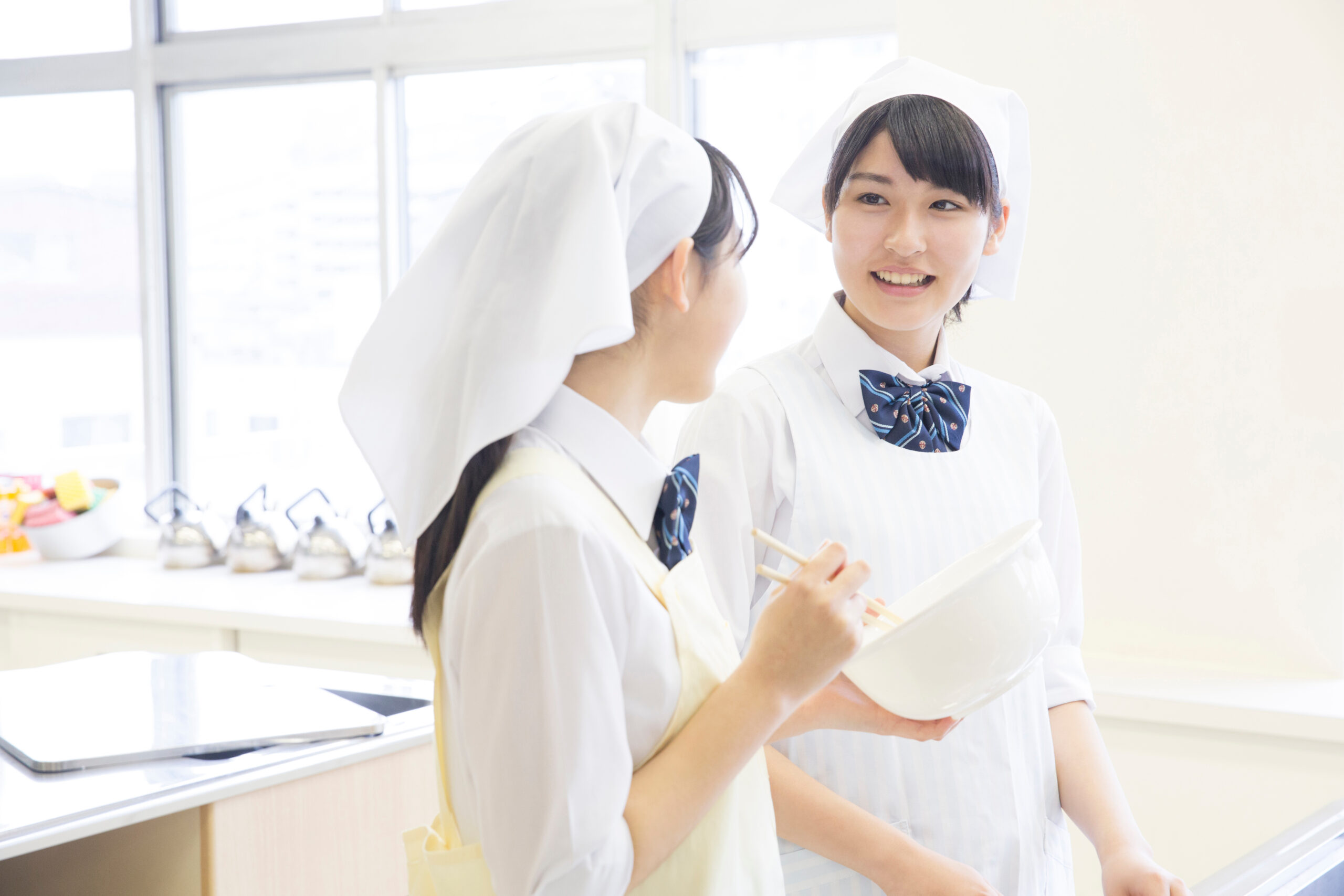 Two young women in white chef uniforms with hats share a conversation in a bright kitchen or classroom setting, one holding a clipboard.