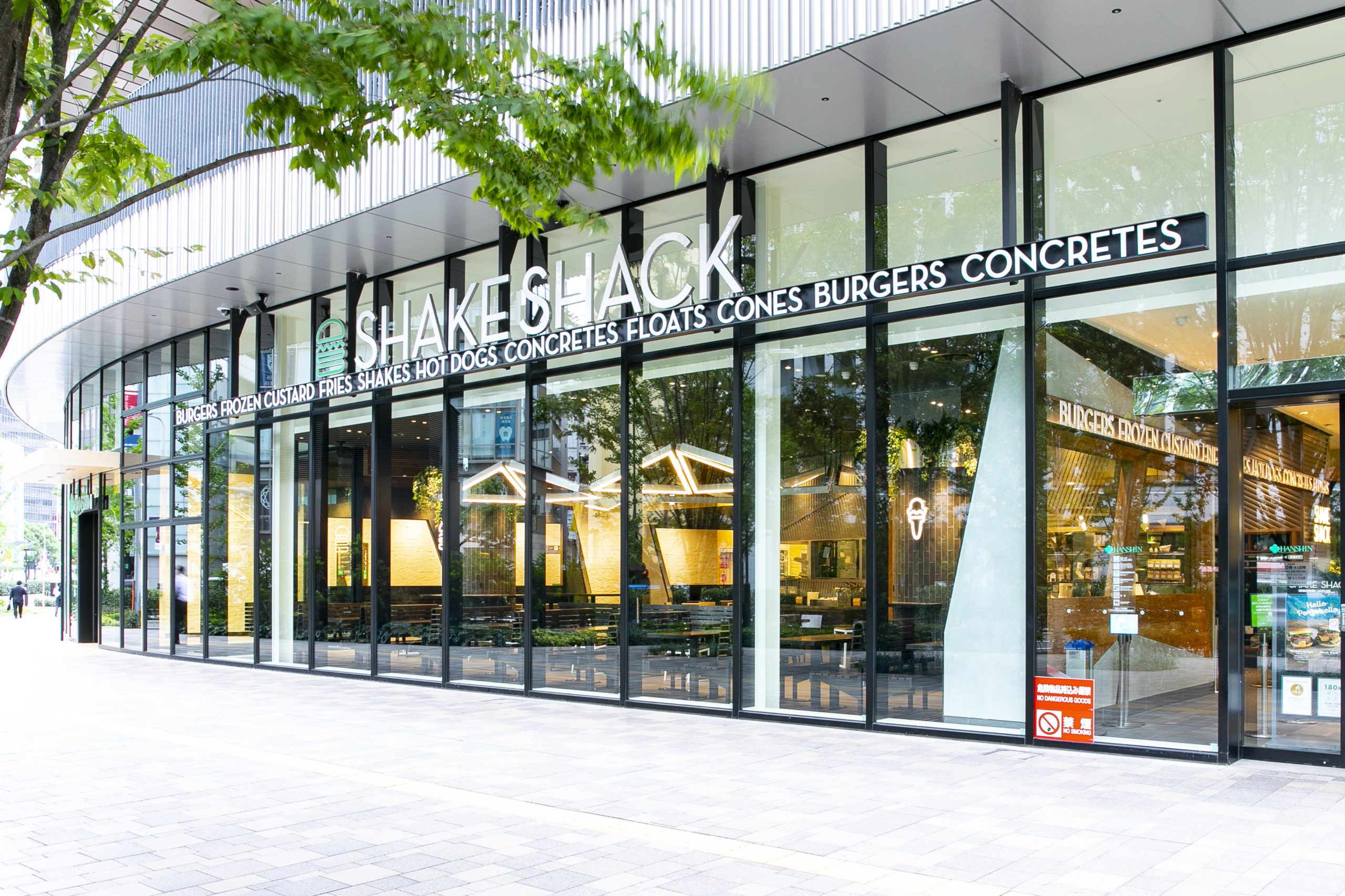 Shake Shack storefront with glass walls, a bold white sign, and green trees in front.