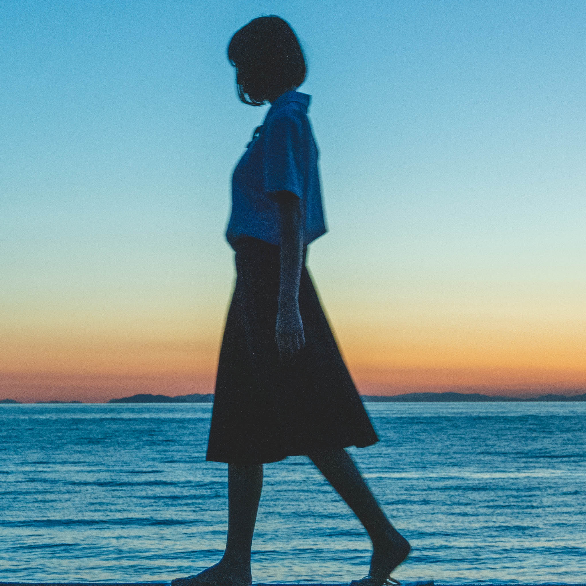 Silhouette of a person walking along the shore at sunset, blue sky fading to light near the horizon.