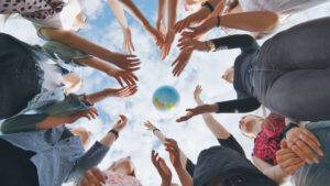 Group of people standing in a circle, hands reaching toward a globe in the center against a cloudy sky.